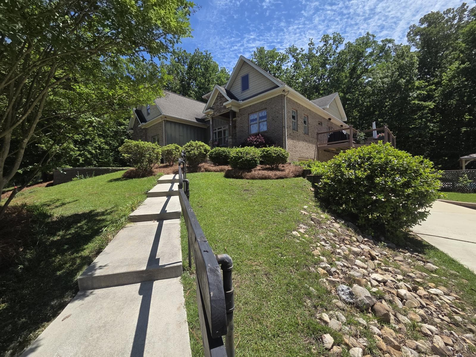 Concrete walkway with black railing leads up a grassy hill toward a large brick house.